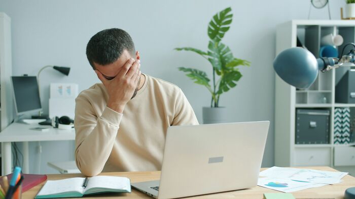 A man in an awkward situation, covering his face with his hand while working on a laptop.