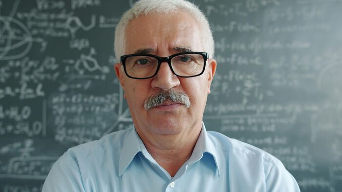 Older male teacher wearing glasses standing in front of a chalkboard in school