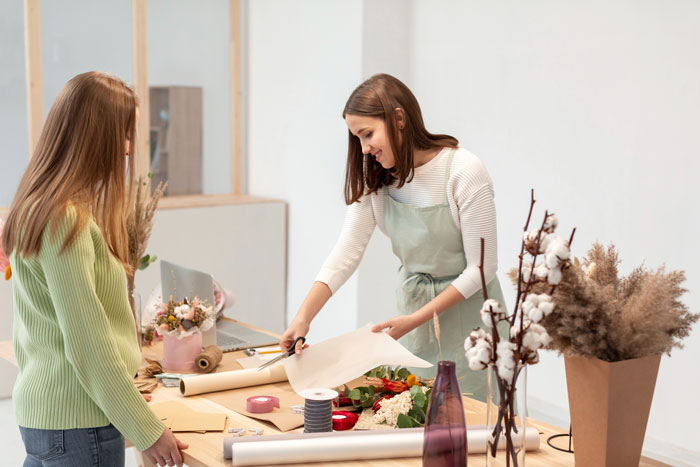 A woman cutting wrapping paper at a table with another woman looking on, surrounded by flowers for a bachelorette party. A woman cutting wrapping paper at a table with another woman looking on, surrounded by flowers for a bachelorette party.