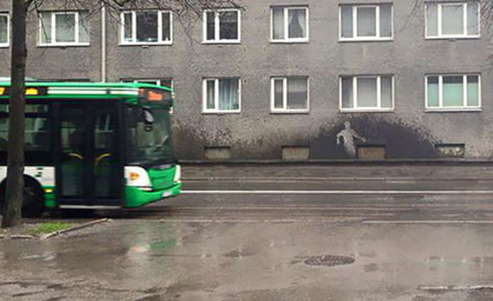 A green bus on a wet street next to a building with a shadow figure on the wall. A creative 'When You See It' visual.