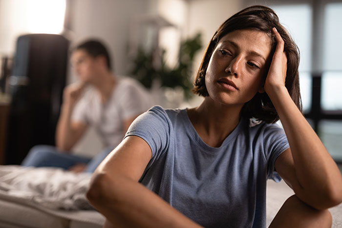 Woman looking stressed and tired while man in background appears distant, showing relationship strain