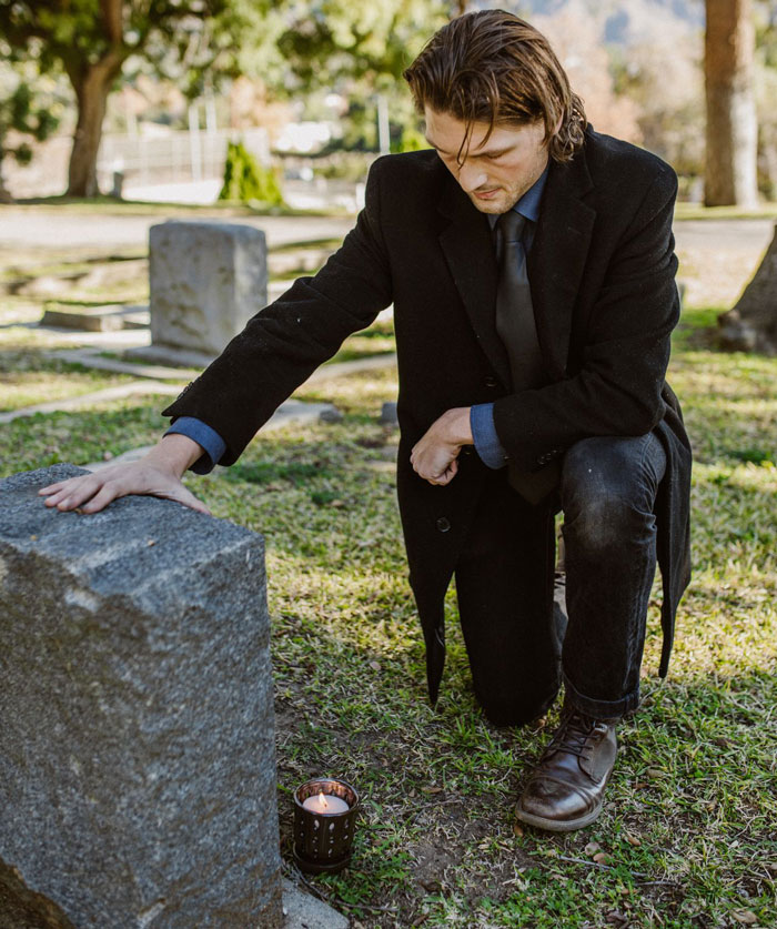 A grieving husband kneels at a tombstone in a cemetery, a candle lit nearby, thinking about his late ex. A grieving husband kneels at a tombstone in a cemetery, a candle lit nearby, thinking about his late ex.