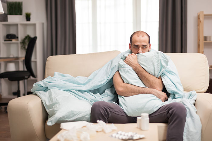 Man wrapped in blanket looking unwell on sofa amid sickness and d***s Man wrapped in blanket looking unwell on sofa amid sickness and d***s
