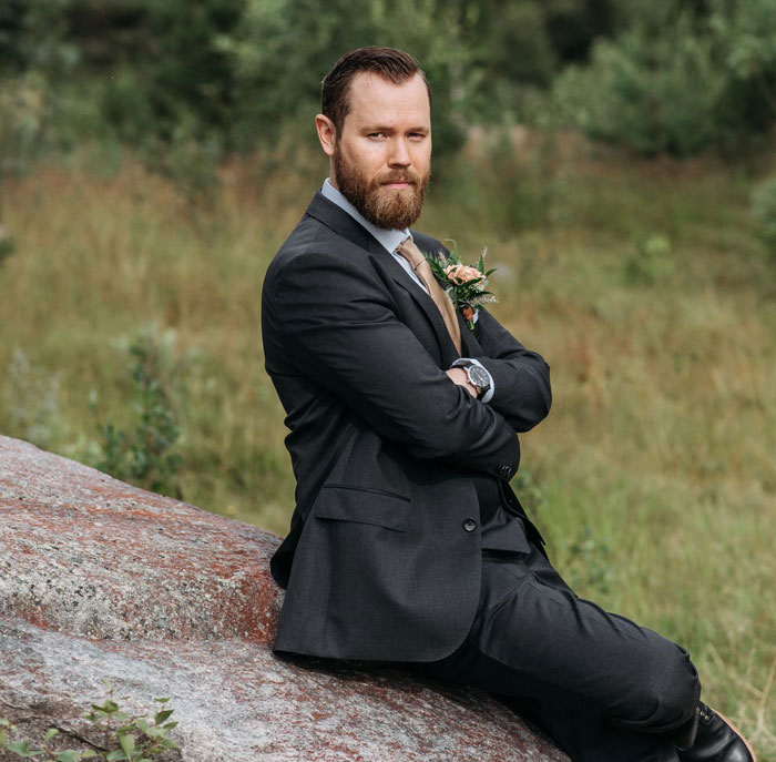 A groom in a dark suit with a beard sits on a rock, looking disgruntled, making his wedding all about him.