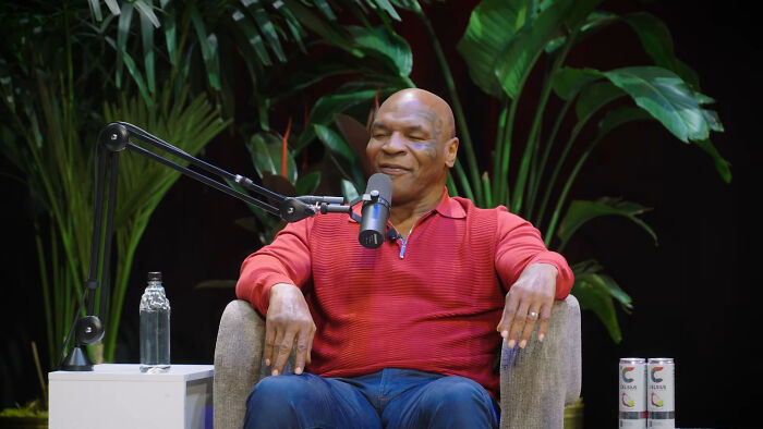 Masked man in red top seated for celebrity encounters podcast discussion