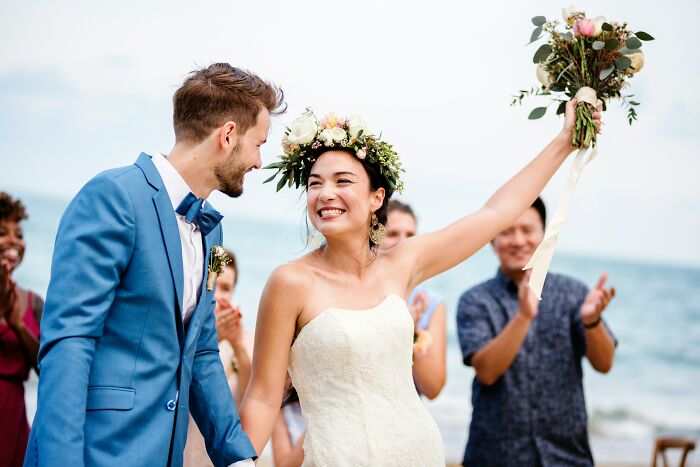 Joyful bride raising bouquet celebrating with groom at beach wedding
