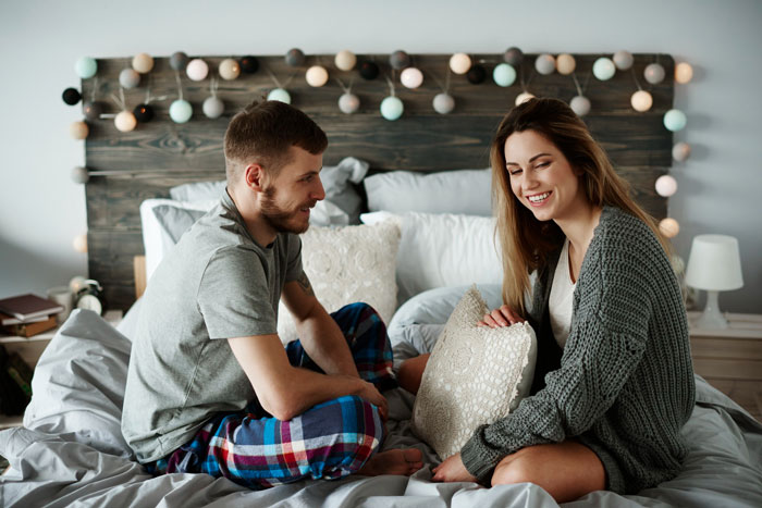 A happy couple, the author and her boyfriend, sit on a bed with decorative lights, discussing his claim of co-writing her book.