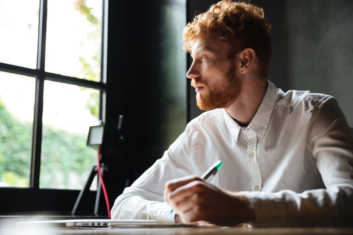 A man with red hair and beard, possibly an author, looking thoughtfully out a window.