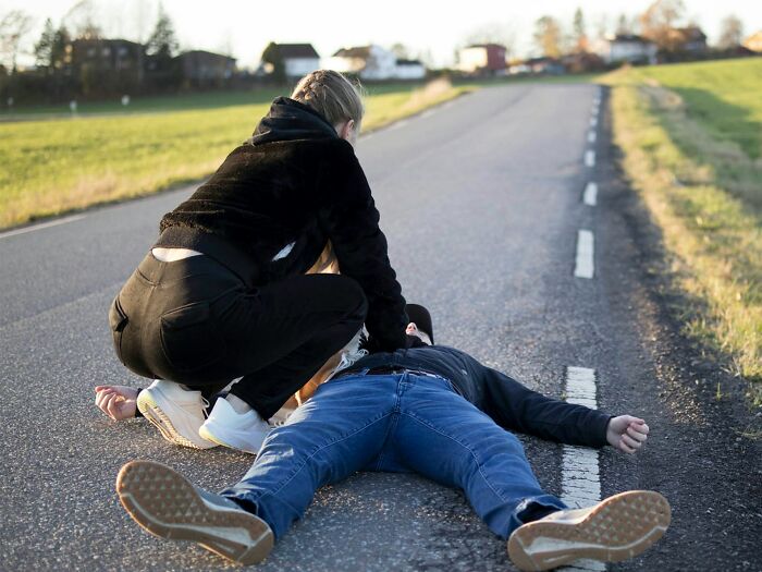 A woman is helping a man lying on the ground in the middle of a road, illustrating something creepy.