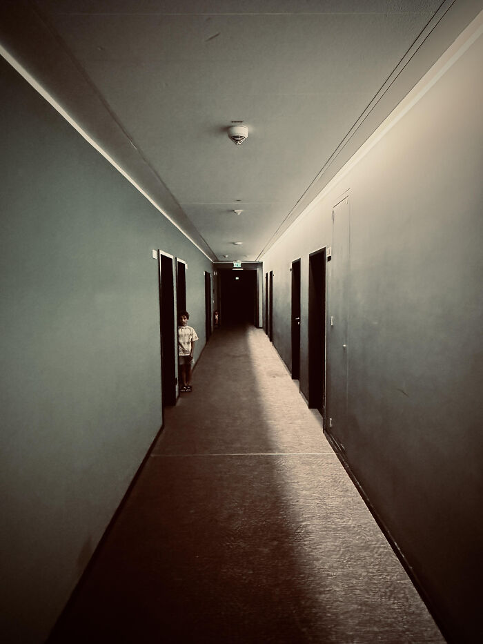 A long, dark, and unsettling hotel hallway, a classic example of liminal spaces, with a child peeking from a door.