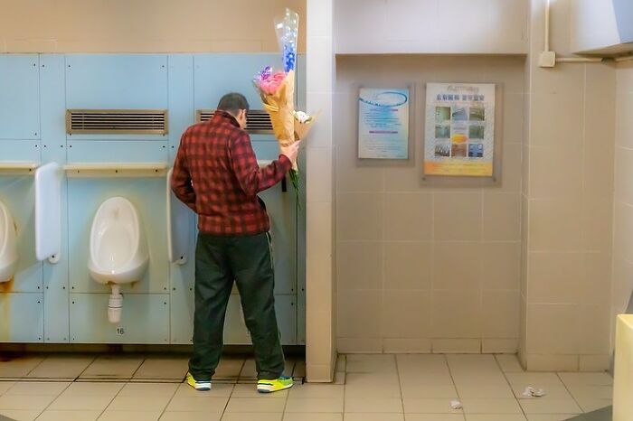 A photographer captures humor in everyday life: a man in a public restroom holding a bouquet of flowers.