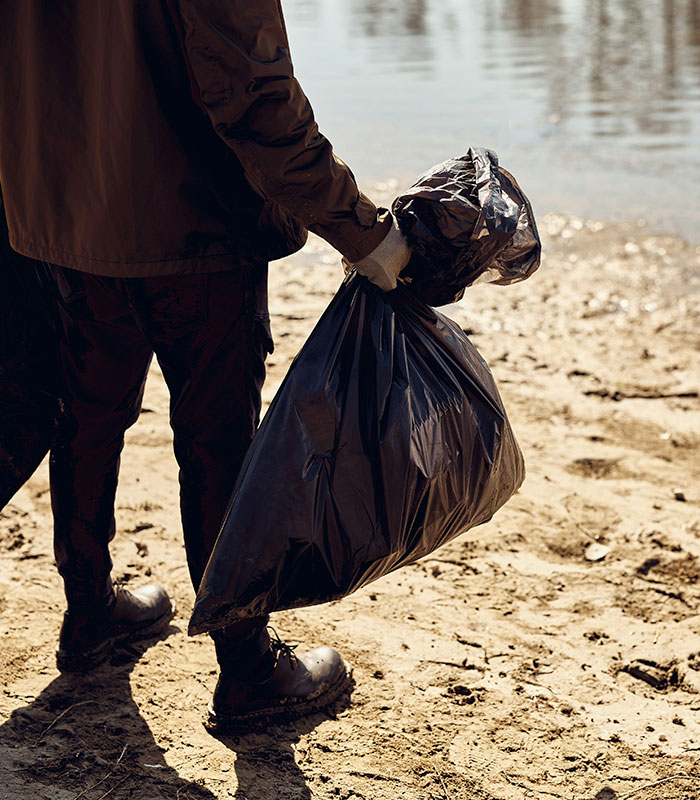 A person in brown clothes and boots holding a black trash bag while standing on a sandy riverbank, part of a cleanup. A person in brown clothes and boots holding a black trash bag while standing on a sandy riverbank, part of a cleanup.