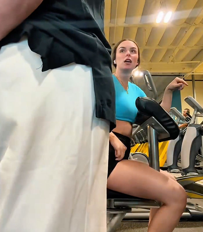 A woman in a blue top and black shorts on a gym machine, looking up at someone out of frame. Gym Karen demands the machine.