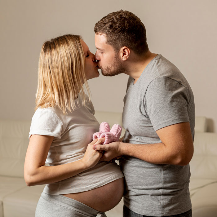 A pregnant woman and her husband kissing, holding baby booties. This image relates to a pregnant wife and a cheater boyfriend.
