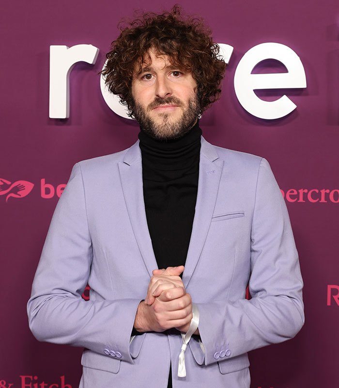 A man with curly hair and a beard, affected by hypospadias, posing in a suit.