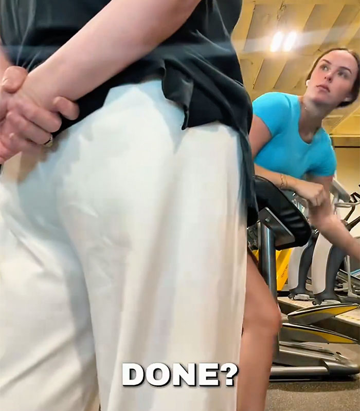 A woman in a blue top and black shorts sitting on a gym machine, looking annoyed. Gym Karen demands to use the machine.