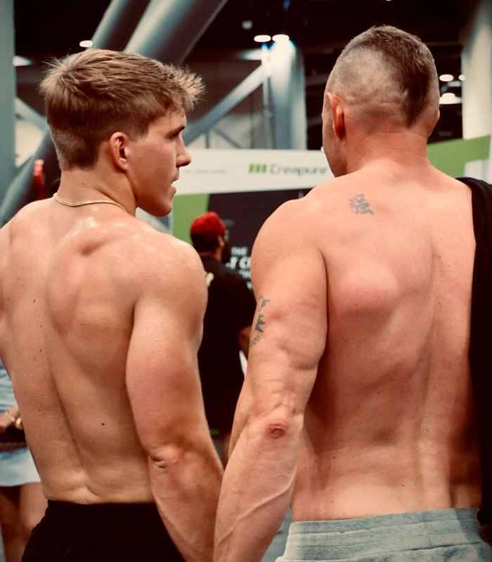 Two muscular men, shirtless, showing their backs in a men-only gym environment.