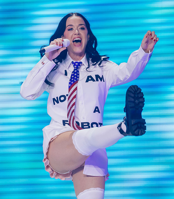 Katy Perry performing in a white shirt and American flag tie, kicking her leg up during a live show.