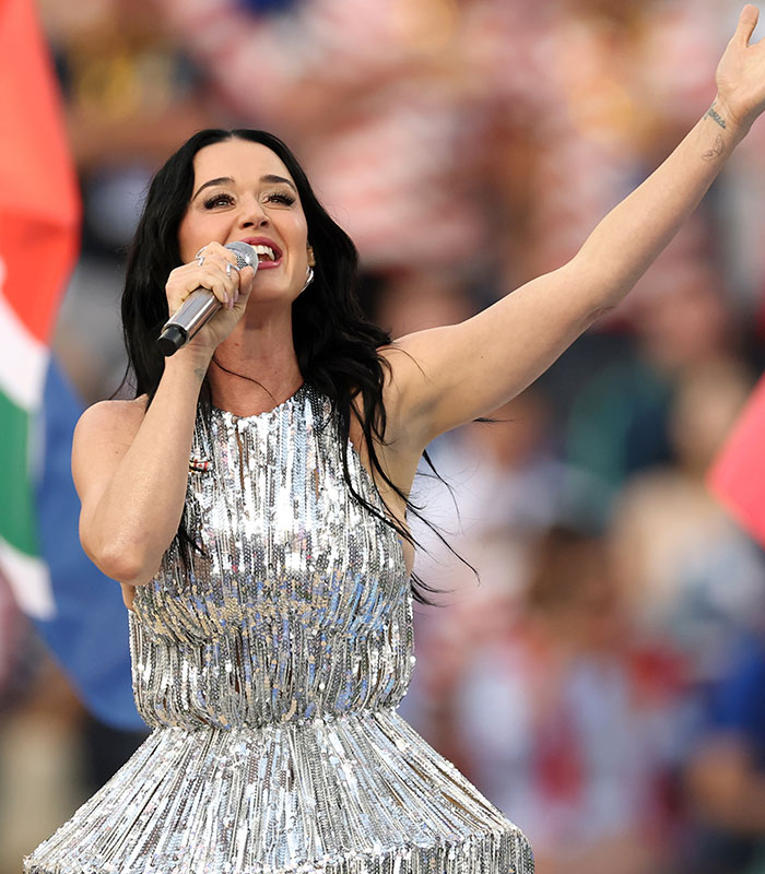 Katy Perry performing live, wearing a shimmering silver outfit. She sings into a microphone with her arm raised, amidst a concert crowd.