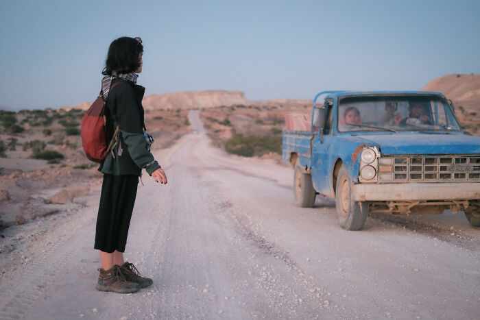 A person with a backpack on a dirt road, next to a blue truck, representing the human side of Strait of Hormuz.