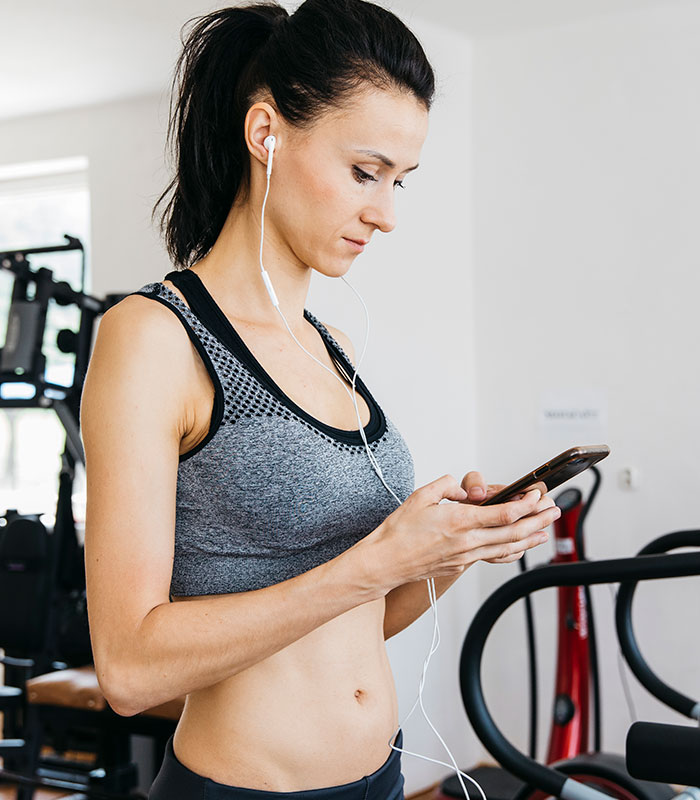 A woman in a sports bra listens to music on wired headphones while checking her phone at the gym, focused on her gym workout.