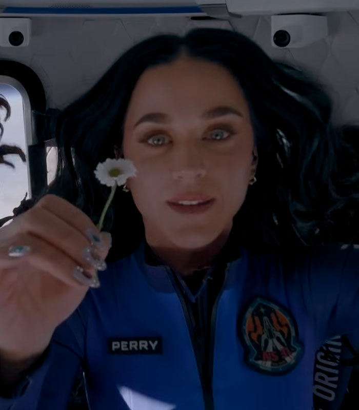 Katy Perry looking at the camera, holding a small white flower, appearing to be in a bottle.