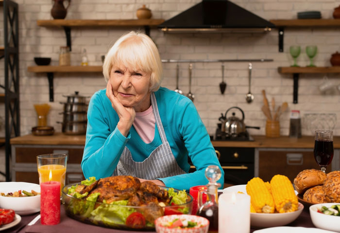 An older woman with blonde hair, wearing a blue top and apron, looking displeased at a Thanksgiving dinner table with essential oil infused holiday dishes. An older woman with blonde hair, wearing a blue top and apron, looking displeased at a Thanksgiving dinner table with essential oil infused holiday dishes.