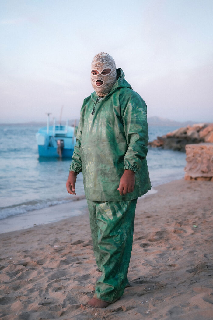 A person in a balaclava and green suit standing on a beach with a boat in the background, a human side of Strait of Hormuz.
