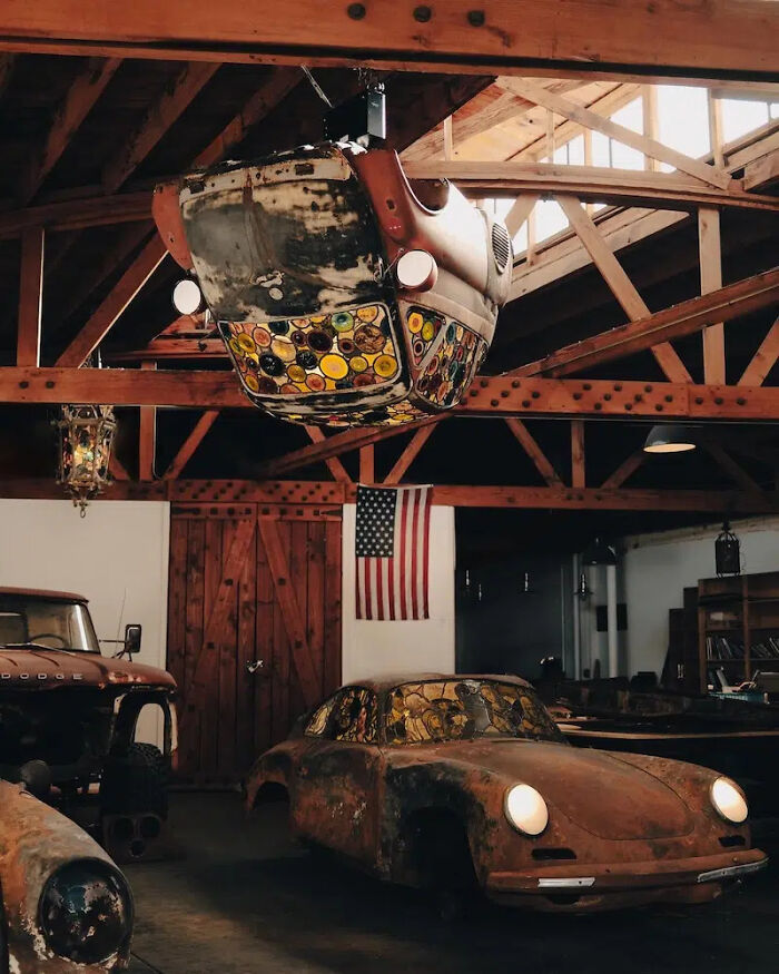 Two burned and rusted Porsche 911 sculptures, one on the ground and one hanging, with stained-glass elements.