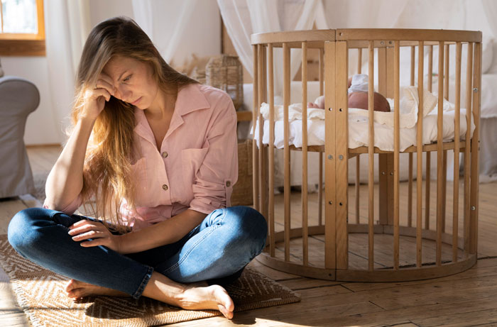 A stressed woman sitting on the floor with a baby in a crib, depicting the struggles of mothers after men walked out.
