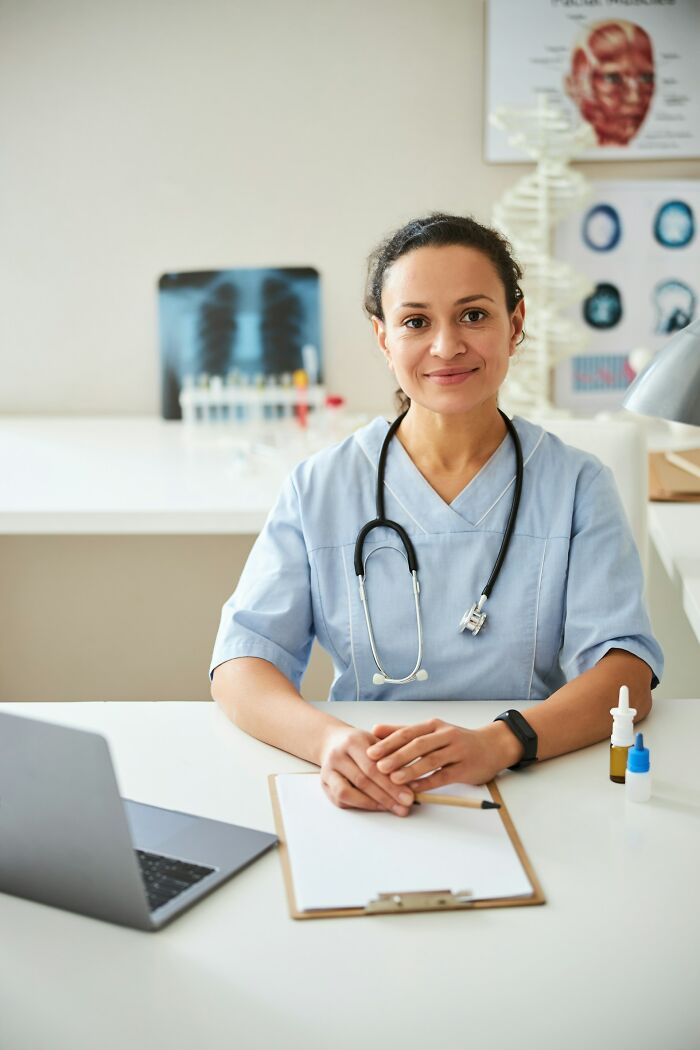 A female doctor with a stethoscope around her neck sits at a desk with a laptop and clipboard, smiling at the camera, highlighting micro feminism clap backs.
