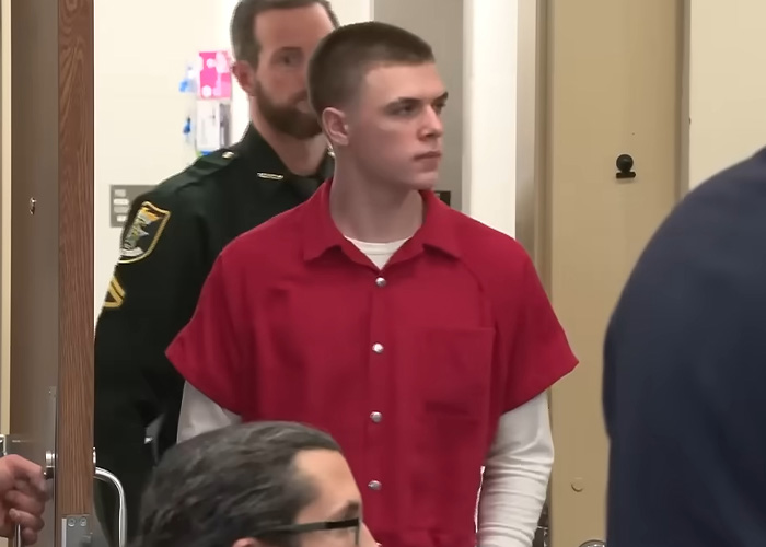 The 18-year-old attacker in a red uniform, escorted by a police officer, stands during his prison sentence.