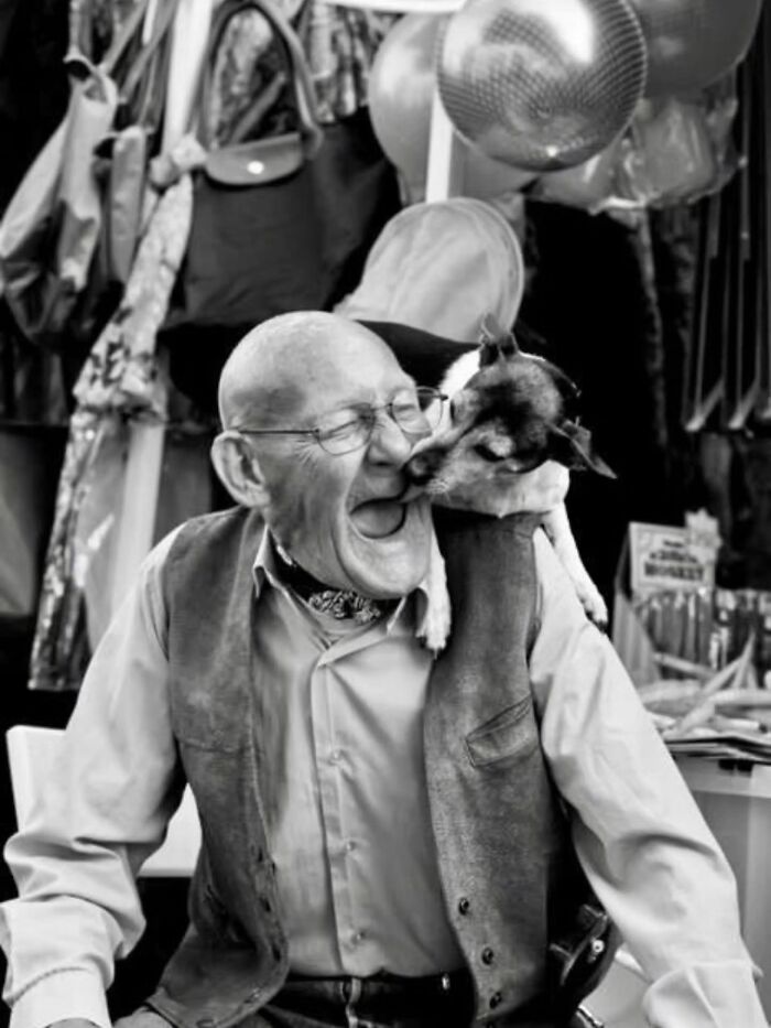 A black and white street photo of an older man laughing while a small dog licks his face, capturing the joy of animals.