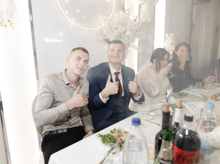 Funny wedding photo of guests giving thumbs up at a table, with the bride and groom in the background.