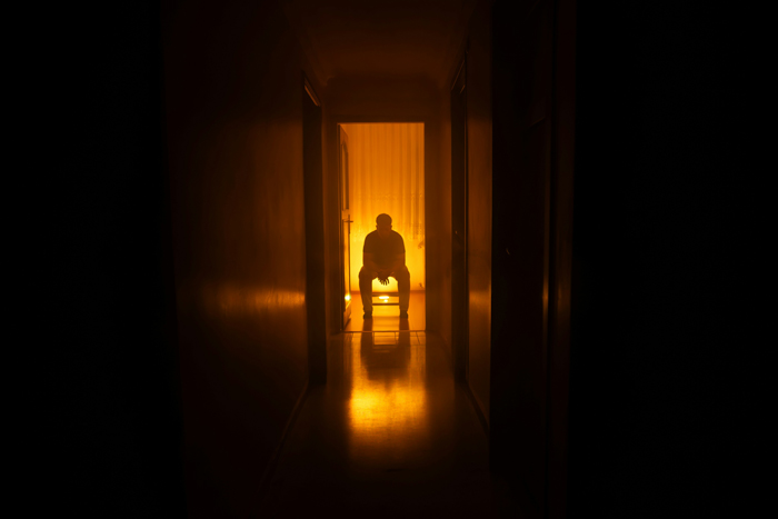 A shadowy figure sits on a chair at the end of a dark, orange-lit hallway, creating a creepy and mysterious scene.