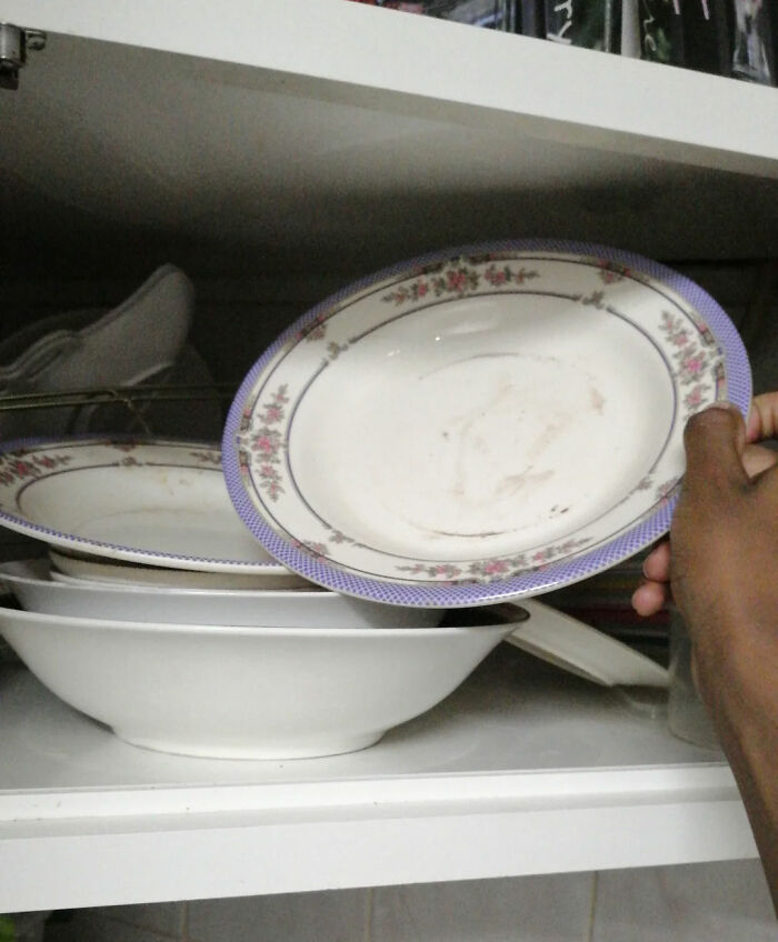 A hand holding a dirty plate with a floral pattern, showing a messy dish cabinet in a woman's home, men freaked out.
