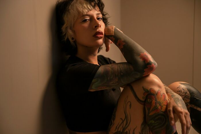 A woman with tattoos on her arms and legs, sitting in a woman's home, looking freaked out.