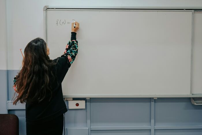 A teacher with long hair writes a math problem on a whiteboard in a classroom, illustrating teachers' work with Gen Alpha.