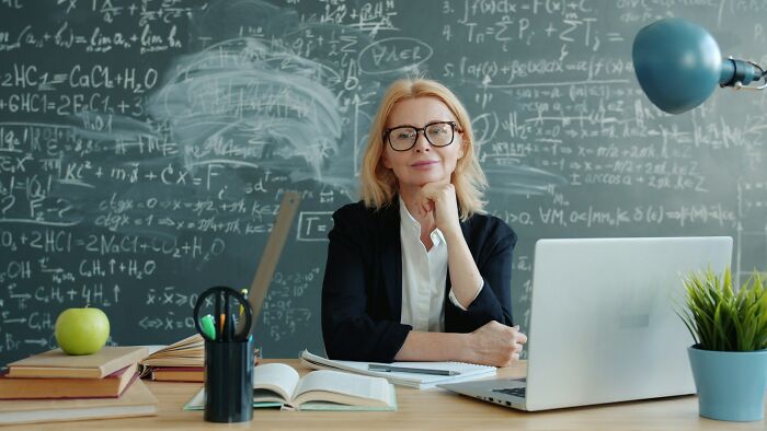 A female teacher smiles at her desk in a classroom with a chalkboard filled with equations, representing teachers' experiences with Gen Alpha.