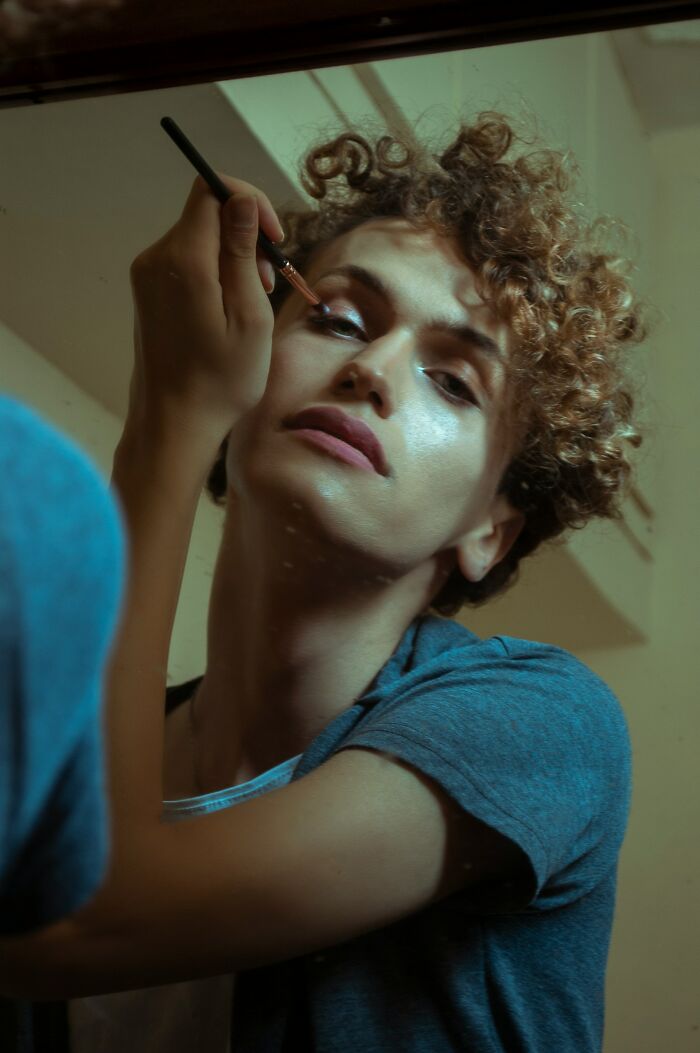 A person with curly hair applying makeup in a mirror, possibly revealing a deep, secret aspect of their identity.
