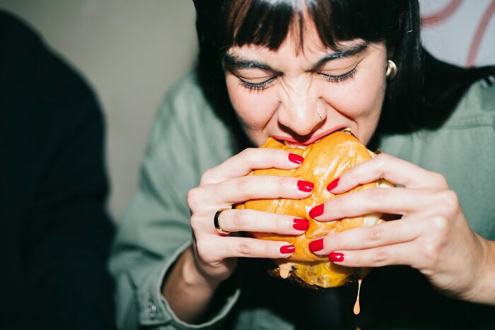 A woman with red nail polish intensely biting into a juicy burger, reflecting a messy, deep secret.