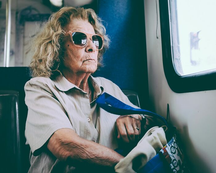 An older woman with sunglasses sits on a train, reflecting on her deep secrets.