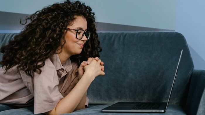 A young woman with curly hair and glasses, lying on a couch and looking at a laptop, contemplating a boyfriend's gift.