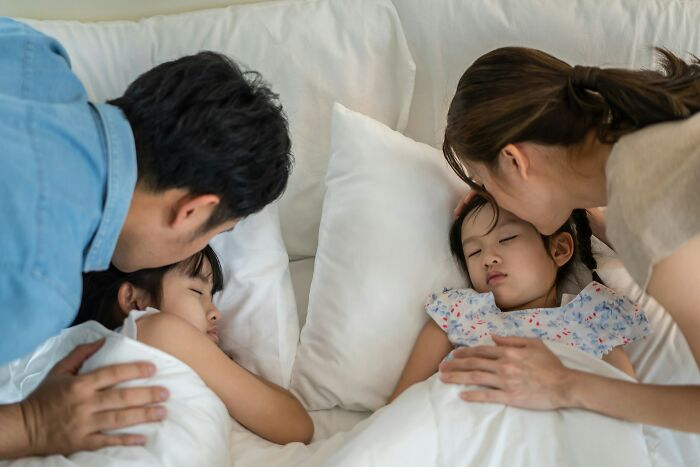 Parents tenderly kissing their sleeping kids in bed, representing kids questions that stump parents.