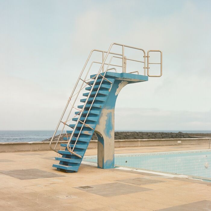 An empty blue diving board with stairs overlooking an abandoned swimming pool and the ocean. reFocus Contest photo capturing the world.