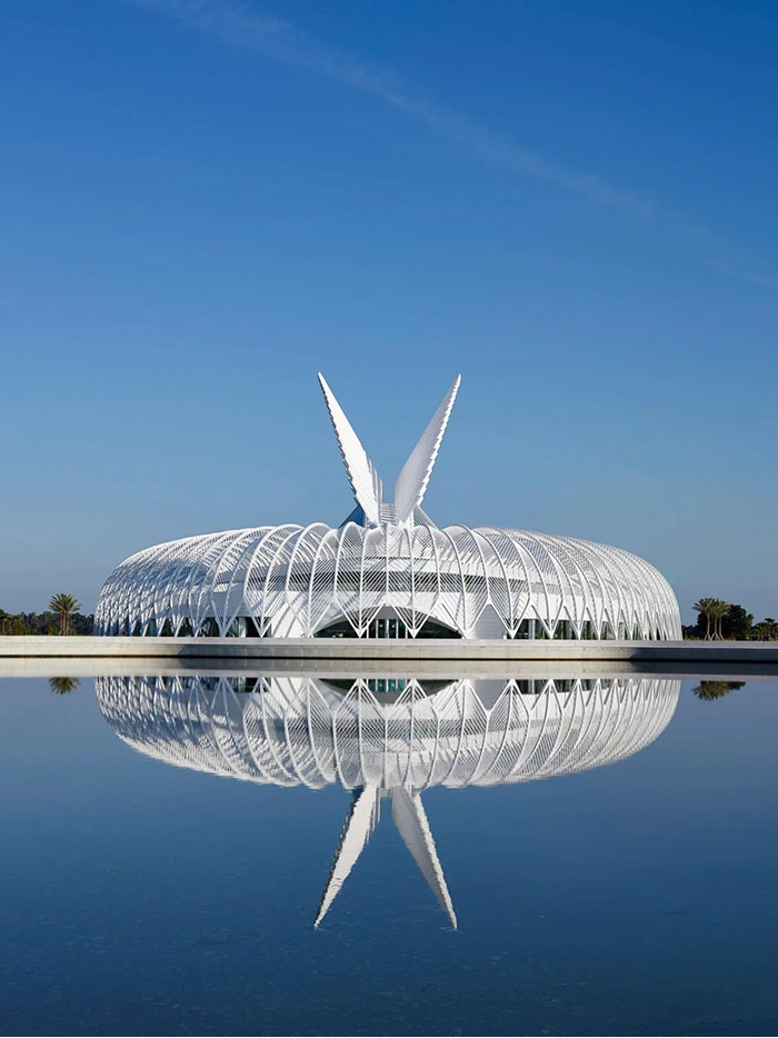 A futuristic modern architecture design of a white, curvilinear building with upward-pointing features, beautifully reflected in a calm body of water.