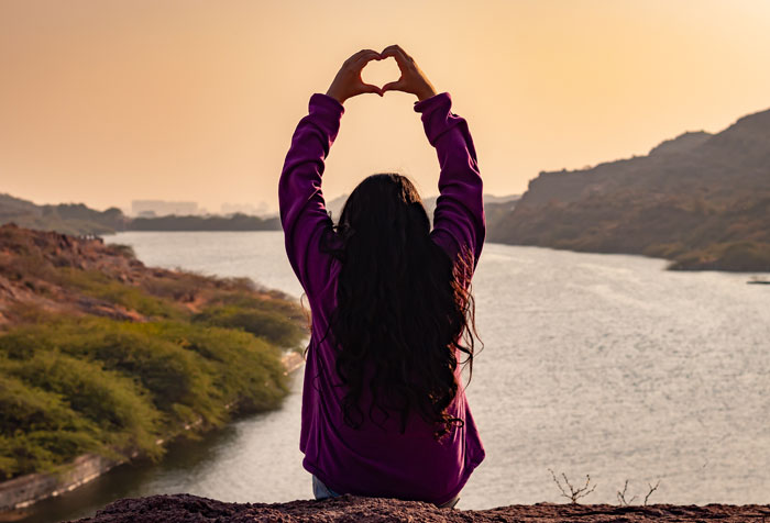 A woman with long dark hair making a heart shape with her hands overlooking a river, embodying hard truths women learned.
