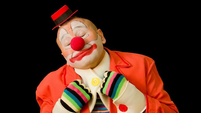 A clown with a red nose and small top hat, wearing an orange jacket and colorful striped gloves, looking pleased with a boyfriend's gift.