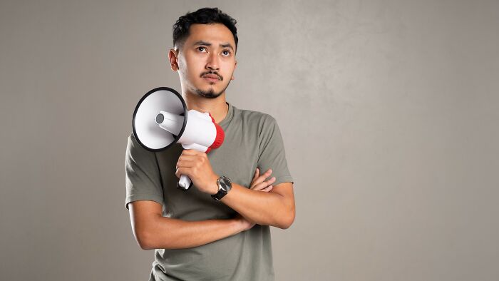 A man with a serious expression, holding a megaphone and looking upwards, with his arms crossed.