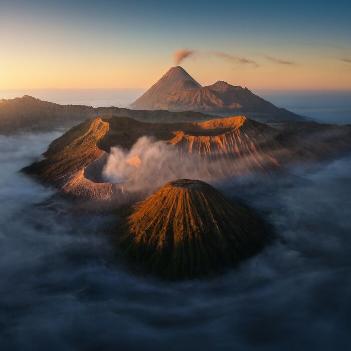An aerial view of multiple volcanic peaks, some with smoke plumes, surrounded by clouds at sunrise. reFocus Contest photo.